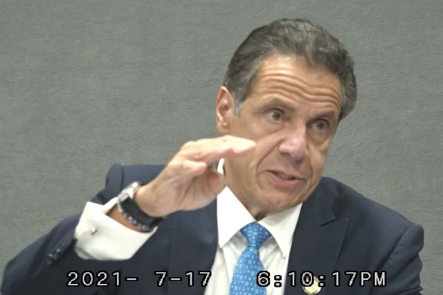 Governor Andrew CUomo, gesturing while speaking and wearing dark suit with light blue tie, in front of drab grey background